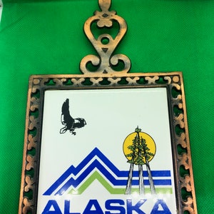 May include: A decorative trivet featuring a white tile with a graphic of Alaska's mountains, trees, and a flying bird, framed in a bronze-colored metal. The word "ALASKA" is printed in blue.
