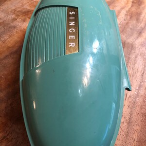 May include: A vintage teal Singer sewing machine cover. The cover has a streamlined, oval shape with a textured side panel. A gold rectangular plate with the brand name "SINGER" is visible on the front.