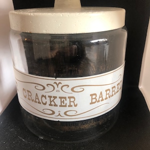 Puede incluir: Un tarro de cristal transparente con tapa blanca y etiqueta dorada con la inscripción "Cracker Barrel". El tarro está lleno de una sustancia oscura, posiblemente galletas o galletas. El tarro está diseñado para el almacenamiento.