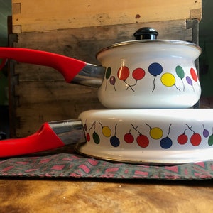 May include: White enamel cookware set with red handles and a black lid. The set features a colorful balloon design. The pot and pan are stacked on a patterned mat, with a wooden background.