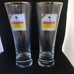 May include: A pair of clear, tall beer glasses with silver rims. Each glass features a label with "ISENBECK PREMIUM" in red and yellow. The glasses are tapered and have a rounded base.