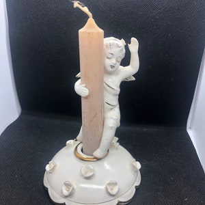 Może przedstawiać: Biały porcelanowy świecznik z cherubinkiem trzymającym beżową świecę. Podstawa jest biała ze złotymi obramowaniami i małymi białymi kwiatowymi akcentami. Świeca ma około 10 cm wysokości.