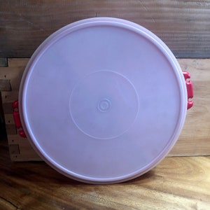 Puede incluir: Tapa de recipiente de plástico rosa claro, de forma circular, con un borde elevado y dos asas de cierre rojas. La tapa tiene un diseño circular en el centro y probablemente es para guardar alimentos. La tapa está sobre una superficie de madera.
