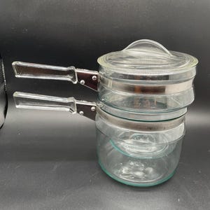 May include: A clear glass double boiler with a lid and two handles. The pot has a silver band around the middle and a clear glass lid with a handle. The handles are clear glass with a brown metal attachment.