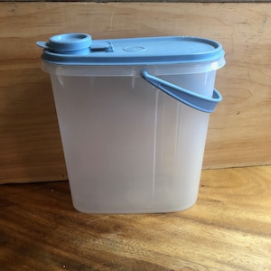 Könnte beinhalten: Ein durchsichtiger Kunststoffbehälter mit hellblauem Deckel und Griff. Der Behälter hat eine rechteckige Form mit abgerundeten Ecken und einem Klappdeckel. Der Griff ist gebogen und an der Seite befestigt. Auf einer Holzoberfläche.