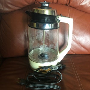 Puede incluir: Cafetera percoladora de vidrio vintage con diseño floral, tapa de acero inoxidable y asa y base color crema. El cable eléctrico es negro. La cafetera está sobre una superficie de cuero marrón.