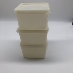 May include: Three off-white, square food storage containers stacked on top of each other. Each container has a matching lid. The containers are made of a durable plastic material. The lids have a slight translucent appearance.
