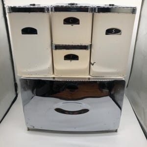 Op de afbeelding: Een vintage keukenkanjesset met een chromen broodtrommel. De set bevat vier crèmekleurige kannen met zwarte labels met de letters "F", "C", "S" en "T". De kannen hebben chromen deksels en sierlijsten. De broodtrommel is chroom.