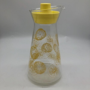 Puede incluir: Una jarra de vidrio transparente con tapa amarilla y boquilla. La jarra está decorada con diseños de rodajas de limón amarillas. La tapa tiene un colador incorporado. Un artículo de cocina vintage.