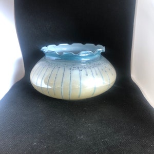 Puede incluir: Un cuenco decorativo de vidrio con un borde con volantes. El cuenco tiene un borde azul claro y un cuerpo color crema con líneas azules verticales y pequeños puntos azules. El cuenco está sobre una superficie negra.