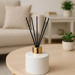 May include: A white ceramic essential oil diffuser with a gold-colored collar, holding several black reeds. The diffuser sits on a light wood table. The background includes a beige sofa and small potted plants, creating a minimalist aesthetic.