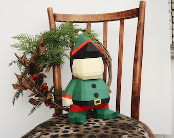 Puede incluir: Una figurita de elfo de papel con un atuendo verde y rojo, sentada en una silla con un cojín de asiento de leopardo marrón. El elfo lleva un gorro rojo y blanco y tiene la cara blanca.