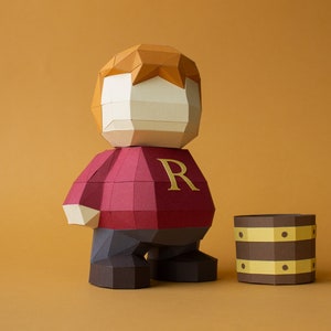 Op de afbeelding: Een papieren figuur van een roodharige jongen die een rode trui draagt met een gouden "R" op de voorkant. De figuur staat naast een bruin papieren vat met gouden accenten.
