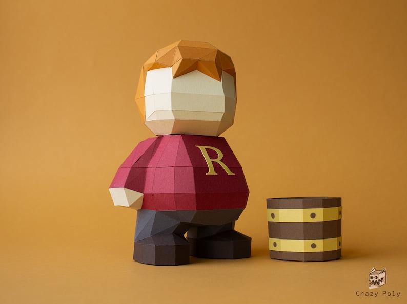 Op de afbeelding: Een papieren figuur van een roodharige jongen die een rode trui draagt met een gouden "R" op de voorkant. De figuur staat naast een bruin papieren vat met gouden accenten.