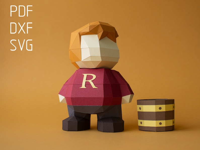 Op de afbeelding: Papiermodel van een roodharig personage dat een rood shirt draagt met een gouden "R" op de voorkant. Het personage staat naast een bruin vat met gouden banden.