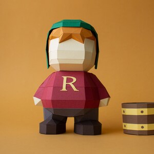 Op de afbeelding: Een papieren figuur van een personage dat een groene hoed en een rood shirt draagt met de letter "R" erop. De figuur staat naast een bruin papieren vat.