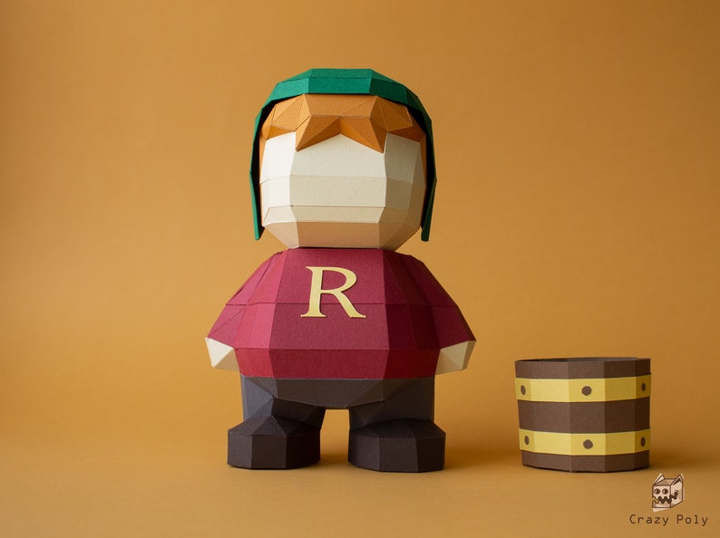 Op de afbeelding: Een papieren figuur van een personage dat een groene hoed en een rood shirt draagt met de letter "R" erop. De figuur staat naast een bruin papieren vat.
