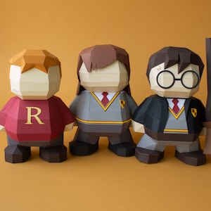 Op de afbeelding: Drie papieren figuren van Harry Potter-personages: Ron Wemel, Hermelien Griffel en Harry Potter. Ron draagt een rode trui met een grote 'R' op de voorkant. Hermelien draagt een grijze trui met een rode das en het Gryffindor-wapen. Harry draagt een zwarte mantel met een rode das en het Gryffindor-wapen. Hij houdt een bruine en zwarte bezem.