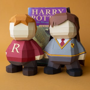 Op de afbeelding: Twee papieren figuren van Harry Potter-personages, Ron Wemel en Harry Potter, die naast een exemplaar van het boek "Harry Potter en de Gevangene van Azkaban" van J.K. Rowling staan. Ron draagt een rood-gouden Gryffindor-uniform met de letter "R" op zijn borst. Harry draagt een grijs-zwart Hogwarts-uniform met een rode stropdas en het Hogwarts-wapen op zijn borst.