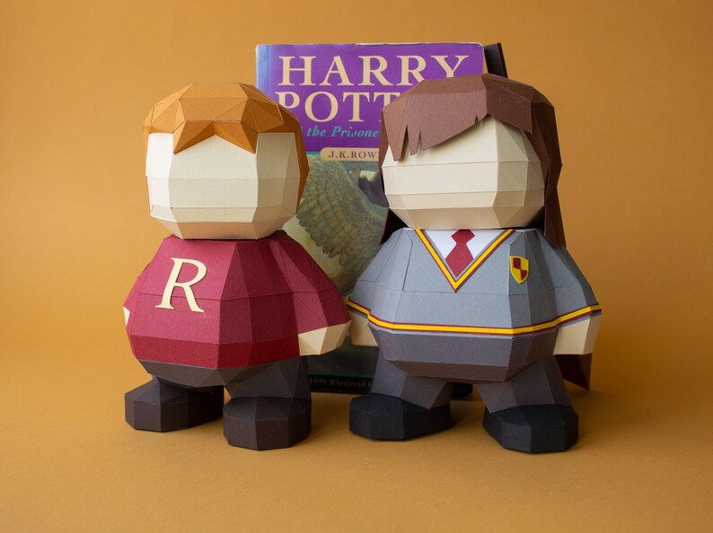 Op de afbeelding: Twee papieren figuren van Harry Potter-personages, Ron Wemel en Harry Potter, die naast een exemplaar van het boek "Harry Potter en de Gevangene van Azkaban" van J.K. Rowling staan. Ron draagt een rood-gouden Gryffindor-uniform met de letter "R" op zijn borst. Harry draagt een grijs-zwart Hogwarts-uniform met een rode stropdas en het Hogwarts-wapen op zijn borst.