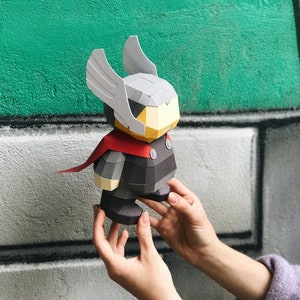 May include: A papercraft figure of Thor, featuring a gray helmet with wings, a red cape, and a black and gray outfit. The figure is held in front of a green and gray background.