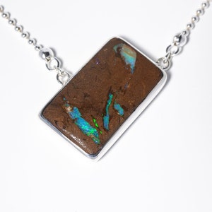 Könnte beinhalten: Ein rechteckiger Boulder-Opal-Anhänger in einer polierten Silberfassung, der an einer Silberkugelkette hängt. Der dunkelbraune Stein zeigt lebendige, irisierende Blitze in Blau und Grün auf seiner texturierten, natürlichen Oberfläche.