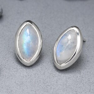 Può includere: Un paio di orecchini a lobo ovali con montatura argentata. Ogni orecchino presenta una pietra iridescente e perlacea con sfumature blu e bianche. Gli orecchini sono esposti su una superficie grigia con accenti di roccia grigia.