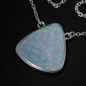 Peut inclure: Un collier en argent avec un pendentif triangulaire orné d'une pierre d'opale blanche. La pierre présente des éclats irisés de lumière bleue, verte et orange. Le pendentif est fixé à une fine chaîne en argent aux maillons circulaires.
