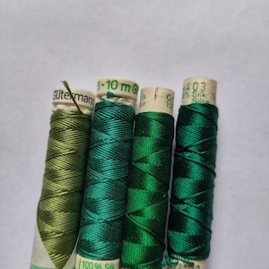 May include: Four spools of silk thread in shades of green. The spools are labeled with the brand "Gütermann" and include details like "100% Silk" and thread numbers. The thread is tightly wound on cardboard spools.