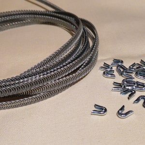 Puede incluir: Una bobina de alambre de metal plateado y un montón de pequeños sujetadores metálicos en forma de U. El alambre está enrollado firmemente y los sujetadores están dispersos cerca. Los artículos están sobre una superficie de color beige claro.