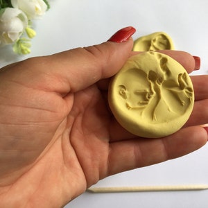 May include: A yellow, circular mold featuring a detailed relief of a baby's profile and a figure. The mold is held in a hand with red nail polish. Other similar molds are visible in the background, along with a wooden stick.