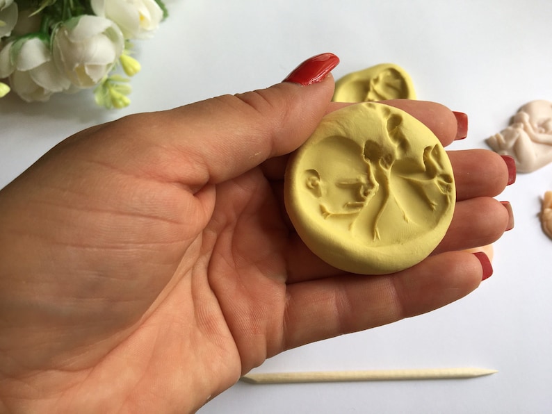 May include: A yellow, circular mold featuring a detailed relief of a baby's profile and a figure. The mold is held in a hand with red nail polish. Other similar molds are visible in the background, along with a wooden stick.