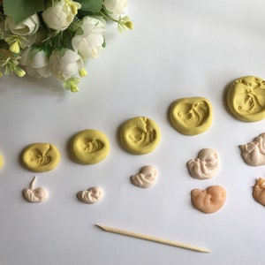May include: A collection of handmade clay sculptures depicting stages of fetal development. The sculptures are in shades of yellow and beige, arranged in a sequence. A wooden tool is also present.