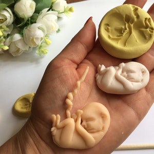 May include: A collection of handmade fetal sculptures and molds. The sculptures are in various stages of development, with a light peach color. A yellow mold with a fetal image is also present. The items are displayed on a white surface.
