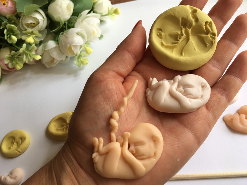 May include: A collection of handmade fetal sculptures and molds. The sculptures are in various stages of development, with a light peach color. A yellow mold with a fetal image is also present. The items are displayed on a white surface.