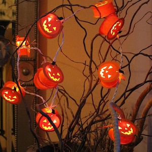 Op de afbeelding: Een lichtsnoer met oranje pompoenlampjes met uitgesneden gezichten, opgehangen aan een witte draad. De lampjes zijn gerangschikt tussen kale, bruine takken, wat een Halloween-sfeer creëert. De pompoenen zijn van binnenuit verlicht.