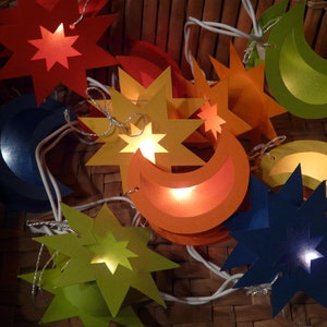 Könnte beinhalten: Eine Lichterkette aus Papier in Form von Sternen und Mondsicheln. Die Lichter sind in verschiedenen Farben, darunter Blau, Grün, Gelb, Orange und Rot. Die Lichter sind mit weißem Draht miteinander verbunden und leuchten.