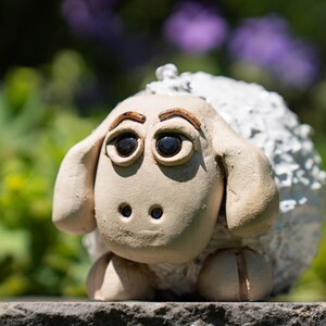 Könnte beinhalten: Eine kleine, handgeformte Keramikfigur eines Schafs mit glattem, hellbeigem Gesicht, dunkelbraun bemalten Augen und geschwungenen Augenbrauen. Der Körper ist mit einer strukturierten, weißen, wolleartigen Oberfläche bedeckt und steht auf zwei Hufen.
