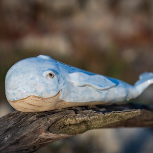 Könnte beinhalten: Eine Keramik-Walfigur mit einer gesprenkelten hellblauen und weißen Glasur auf dem Oberkörper und einer glatten, unglasierten beigen Unterseite. Der Wal hat einen abgerundeten Kopf, ein sichtbares Auge, Seitenflossen und eine Schwanzflosse.
