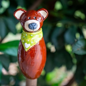 Könnte beinhalten: Eine skurrile, braune Keramik-Bärenfigur mit weiß bespitzten Ohren, einer schwarzen Nase und einem grünen gepunkteten Schal mit einem weißen Herzen. Der Bär ist ein dekorativer Gartenstecker, ca. 15 cm hoch.