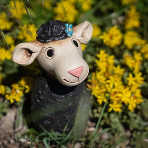 Könnte beinhalten: Eine Keramik-Schaf-Figur mit schwarzem, strukturiertem Körper, cremefarbenem Gesicht und großen runden Augen. Das Schaf hat einen schwarzen Wollschopf auf dem Kopf, der mit einer kleinen blauen Blume verziert ist, sowie eine rosa Nase und abstehende Ohren.