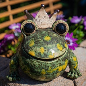 Könnte beinhalten: Keramikfroschfigur mit Krone. Der Frosch ist grün mit gelben Punkten und großen schwarzen Augen. Die Krone ist beige mit dekorativen Details. Der Frosch sitzt auf einer Steinoberfläche, mit lila Blumen im Hintergrund.