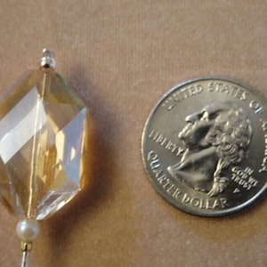 May include: A faceted, amber-colored crystal bead with a silver-colored top and a small pearl bead at the bottom. A U.S. quarter coin is next to the bead for size comparison. The bead has a geometric shape and is likely for jewelry making.