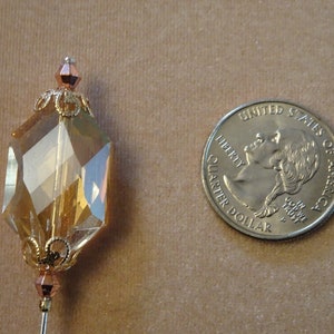 May include: A decorative pin with a large, faceted, amber-colored crystal pendant. The crystal is an elongated hexagon shape, with gold-colored filigree and bead accents. A U.S. quarter coin is next to the pin for size comparison.
