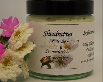 Peut inclure: Un pot blanc avec un couvercle noir et une étiquette blanche qui dit "Shea Butter - White Tea - die natürliche Körperpflege". Le pot est posé sur une surface blanche avec des fleurs blanches et roses.