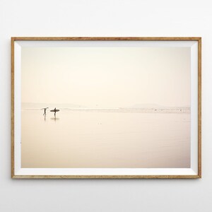 May include: A framed photograph of two people walking on a sandy beach with surfboards. The beach is empty and the sky is a soft, pale pink.