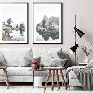 May include: Two framed landscape photographs of a lake and mountains with a reflection of the scenery in the water. The photos are hanging on a white wall above a gray couch with pillows and a throw blanket.
