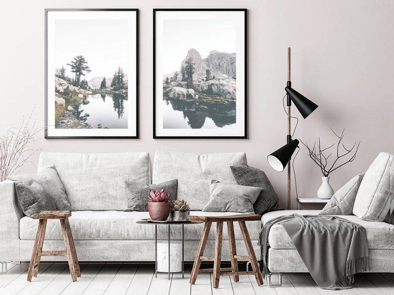 May include: Two framed landscape photographs of a lake and mountains with a reflection of the scenery in the water. The photos are hanging on a white wall above a gray couch with pillows and a throw blanket.