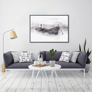 May include: A black framed photograph of a wooden boat on a foggy lake. The boat is tied to a dock with rope. The photo is hanging on a white wall above a gray couch with pillows.