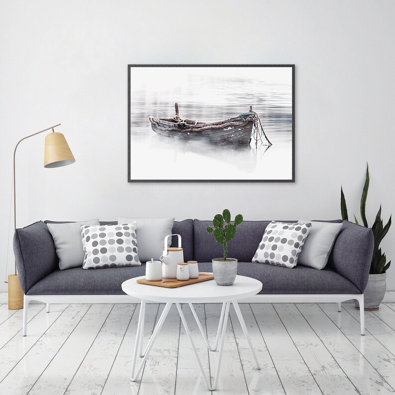 May include: A black framed photograph of a wooden boat on a foggy lake. The boat is tied to a dock with rope. The photo is hanging on a white wall above a gray couch with pillows.